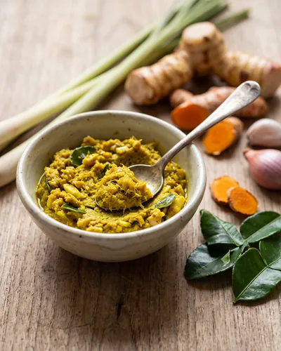Kroeung: a small bowl of fresh, moist, yellow pounded Khmer lemongrass-and-turmeric aromatic paste with a spoon, and its raw ingredients arranged beside the bowl.