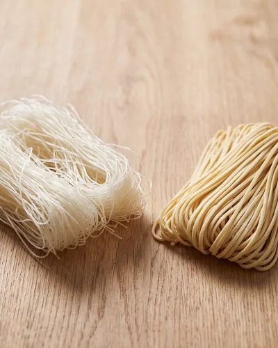 A nest of thin translucent-white dried rice noodles beside a bundle of pale straw-yellow dried wheat noodles on wood, showing the difference in strand width and colour.