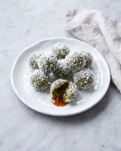 Ondeh-ondeh: small deep-green glutinous rice balls fully rolled in white grated coconut, on a white plate, with one broken open to show the dark gula Melaka palm sugar syrup running out.